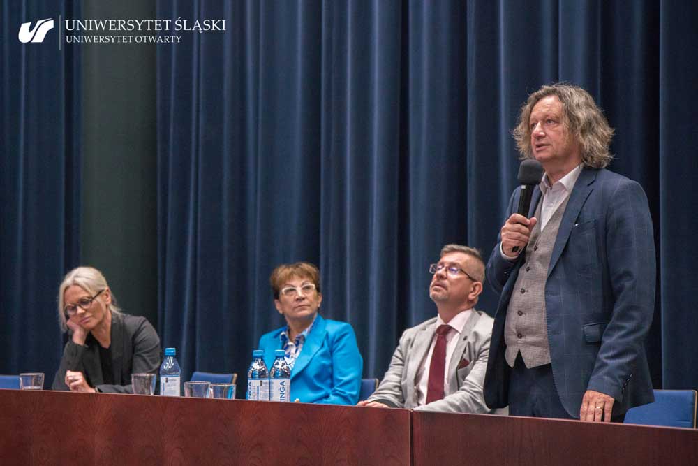 Prof. dr hab. Ryszard Koziołek, rektor Uniwersytetu Śląskiego, stoi po lewej stronie zdjęcia i przemawia, trzymając mikrofon. Obok niego siedzą trzy osoby.