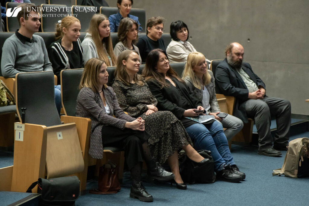 Publiczność siedząca w auli. W pierwszym rzędzie siedzą cztery promiennie uśmiechające się kobiety oraz zamyślony mężczyzna. Za nimi siedzą młode osoby, również uśmiechające się.