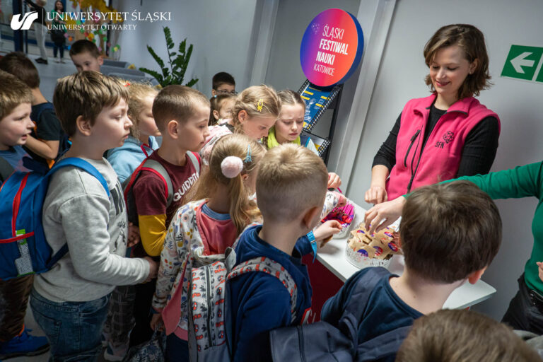 Kobieta w różowej kamizelce z logo Śląskiego Festiwalu Nauki Katowice rozdaje zgromadzonym przy ladzie dzieciom gadżety.