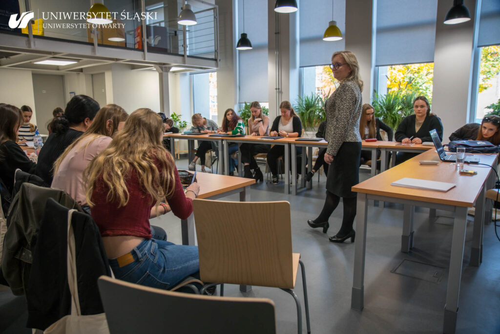 Osoby uczestniczące w Edukatorium pedagogiczno-psychologicznym siedzą przy złączonych w literę U biurkach. Na środku sali stoi kobieta. Patrzy się na jedną z osób.