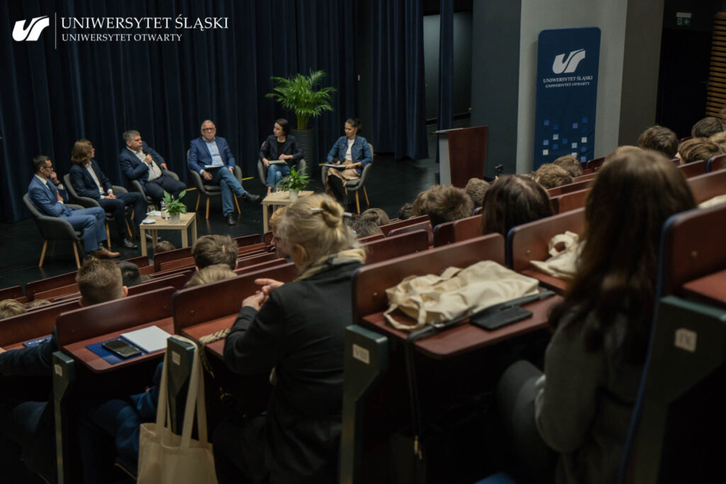 Zdjęcie zrobione z lewej strony i z góry. Tyłem do kamery siedzi publiczność w auli. Na scenie siedzą osoby biorące udział w debacie w ramach Edukatorium obywatelskiego.